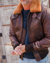 A person wearing a brown leather jacket with a fur collar and blue sweater stands in front of a wooden gate.