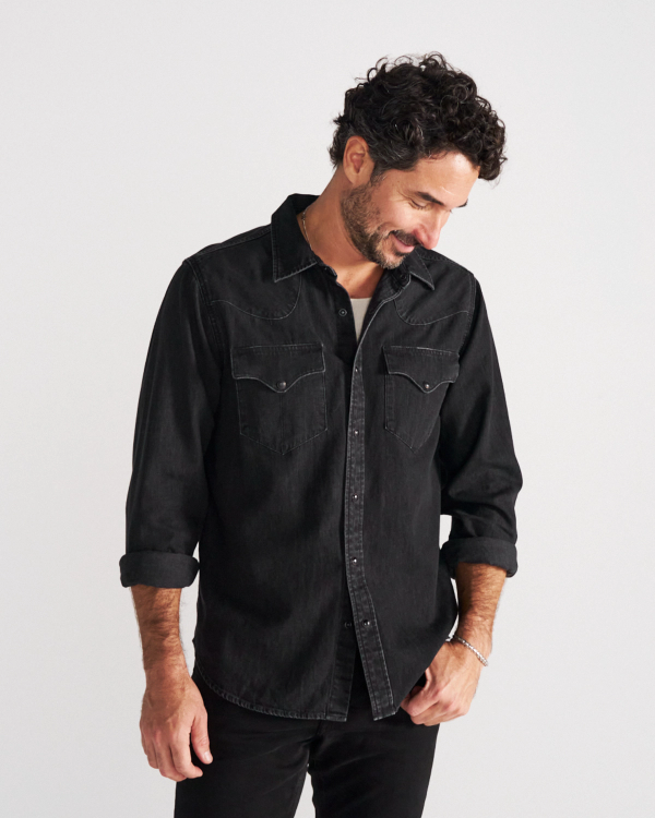 Man with curly hair and a beard, wearing a black denim button-up shirt with rolled sleeves, stands against a plain white background, looking down and slightly smiling.