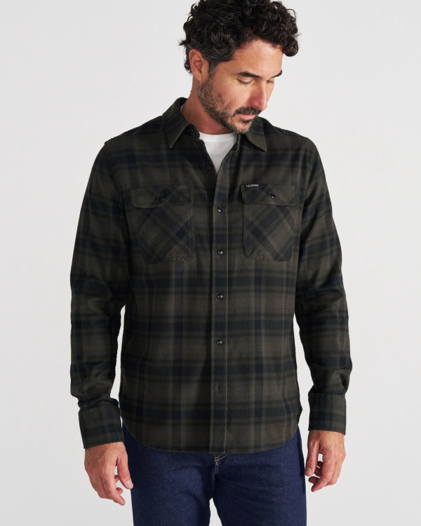 A man stands against a plain background, wearing a dark green and black plaid button-up shirt over a white t-shirt and blue jeans, looking down and to his left.