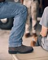 Close-up of a person wearing jeans and brown boots sitting on a vehicle, with another person standing in the background wearing camouflage pants and brown boots.