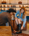 A man assists a woman in trying on black cowboy boots in a store with shelves displaying boots and leather bags in the background.