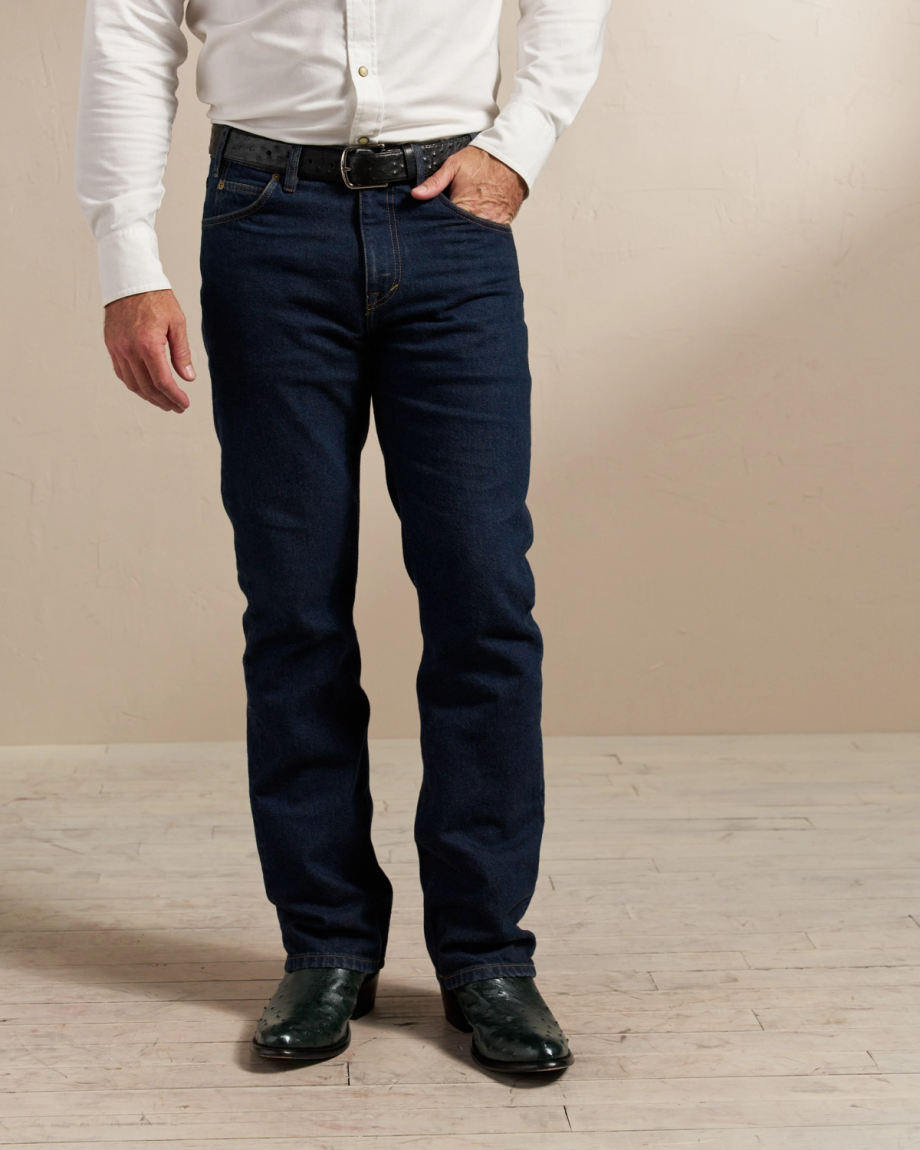 A person wearing dark blue jeans, a white long-sleeved shirt, a black belt, and black shoes stands in front of a beige wall. Only the lower half of their body is visible.