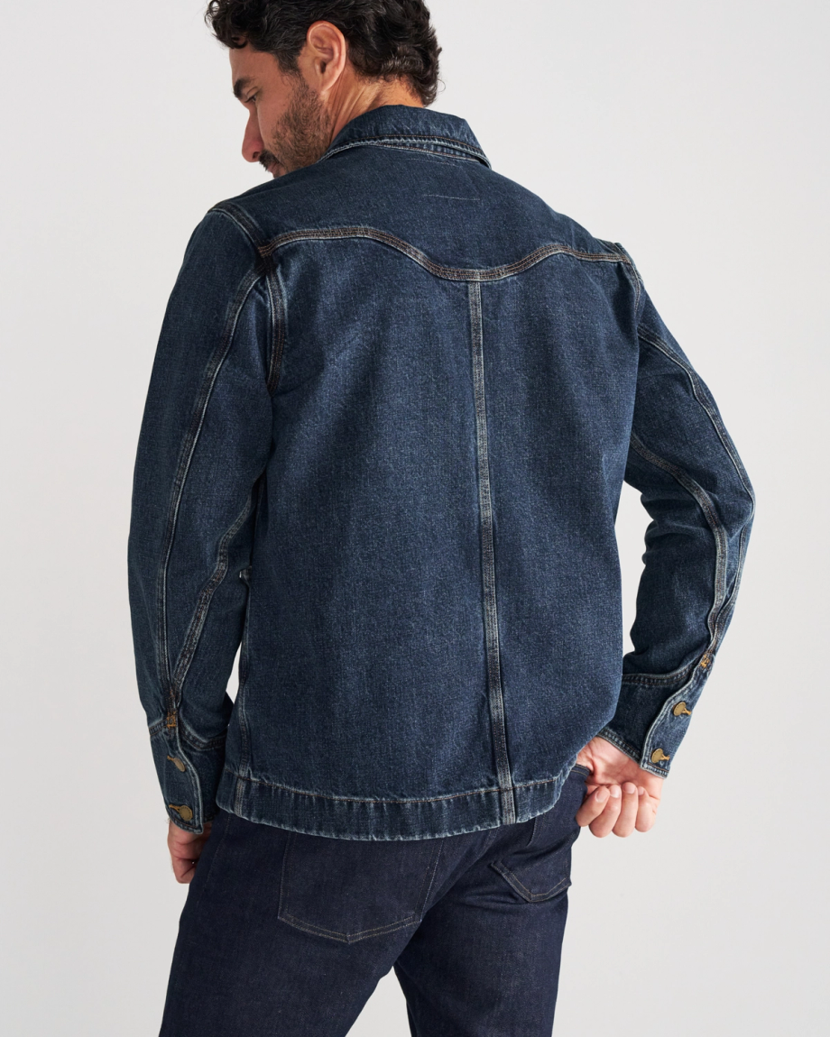 A man wearing a dark blue denim jacket and matching jeans stands facing away from the camera against a plain light background.