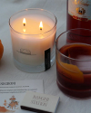 Two lit candles beside a glass of dark beverage with an orange peel, a bottle labeled "Milano," an orange, and a matchbox. Also visible is a card with the word "Negroni" and text below it.