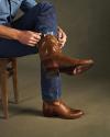A person sitting on a wooden chair pulls on a brown cowboy boot over blue jeans, with another brown boot already on the other foot.