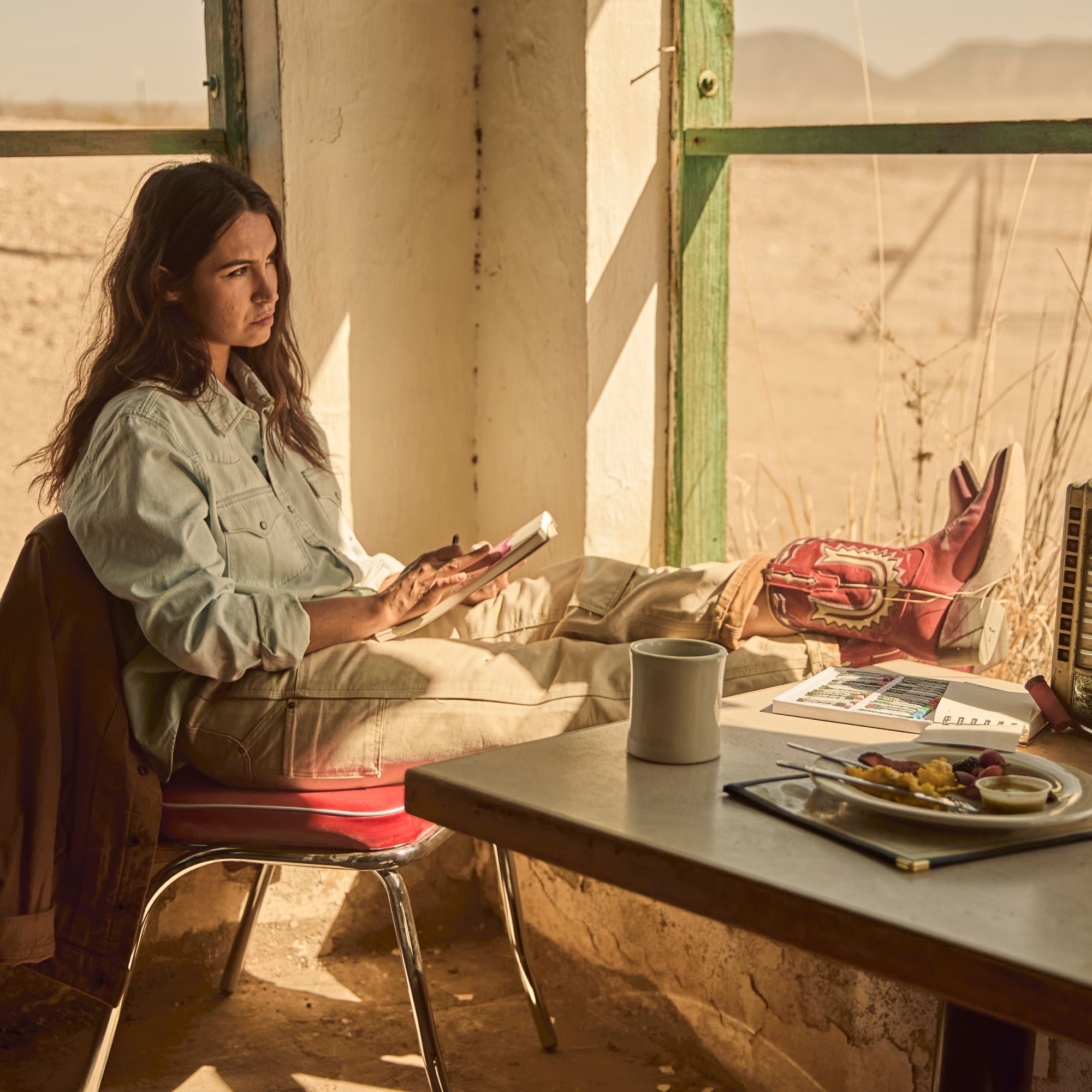 Woman in casual clothes and red boots sits with feet up on a window ledge, holding a phone. A table with food, a mug, and papers is in the foreground. Desert landscape visible outside.