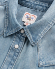 Close-up of a light blue denim shirt with metal snap buttons and a visible brand label reading “Pecos West Western Design.”.