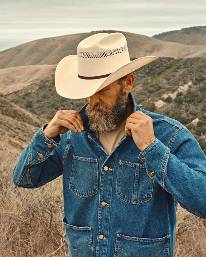 A bearded man adjusts the collar of his denim jacket while wearing a cowboy hat. He stands in a hilly, dry landscape.