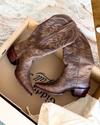 A pair of brown cowboy boots with decorative stitching lies inside a cardboard box on a beige surface.