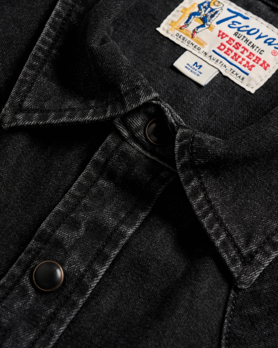 Close-up of a black denim shirt with metal snap buttons and a visible Tecovas Western Denim label on the collar.