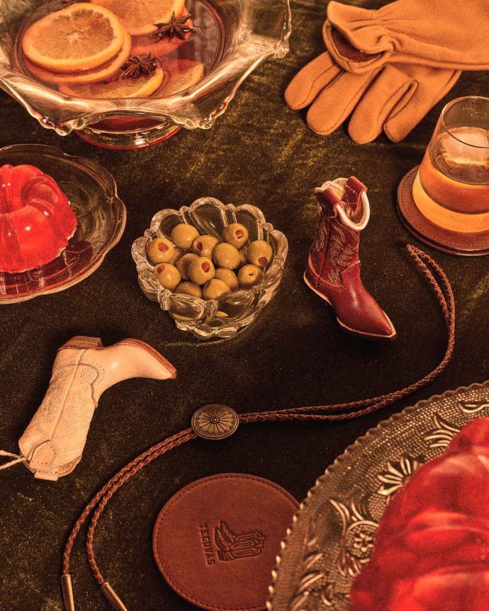 A tabletop with a bowl of olives, a drink, red gelatin, orange slices in punch, leather gloves, two miniature cowboy boots, a bolo tie, and a leather coaster.
