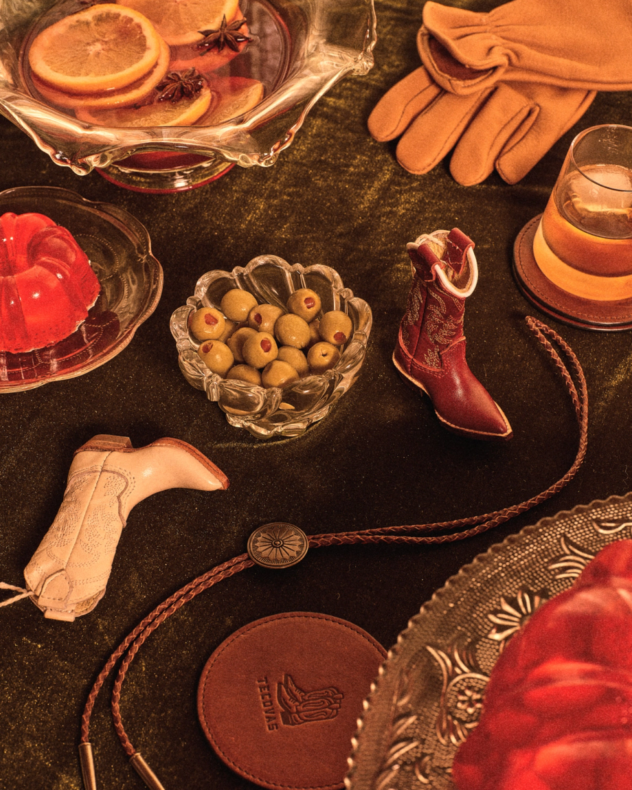 A tabletop with a bowl of olives, a drink, red gelatin, orange slices in punch, leather gloves, two miniature cowboy boots, a bolo tie, and a leather coaster.