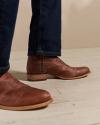 Close-up of a person wearing brown leather boots and dark jeans standing on a light wooden floor.