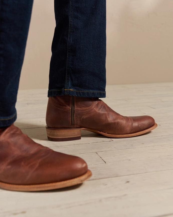 Close-up of a person wearing brown leather boots and dark jeans standing on a light wooden floor.