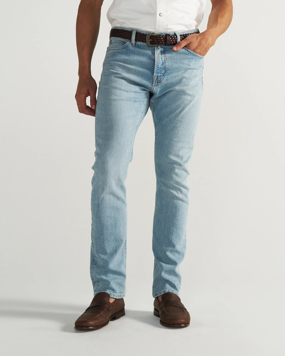 A person wearing light blue jeans, a brown belt, brown shoes, and a white shirt stands against a plain background.