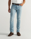 A person wearing light blue jeans, a brown belt, brown shoes, and a white shirt stands against a plain background.