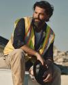 A man in a yellow safety vest and tan pants sits outdoors holding a hard hat, with construction materials and debris in the background.