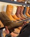 A person holds a brown cowboy boot with an exaggeratedly long toe in a store, with various other cowboy boots displayed on shelves in the background.
