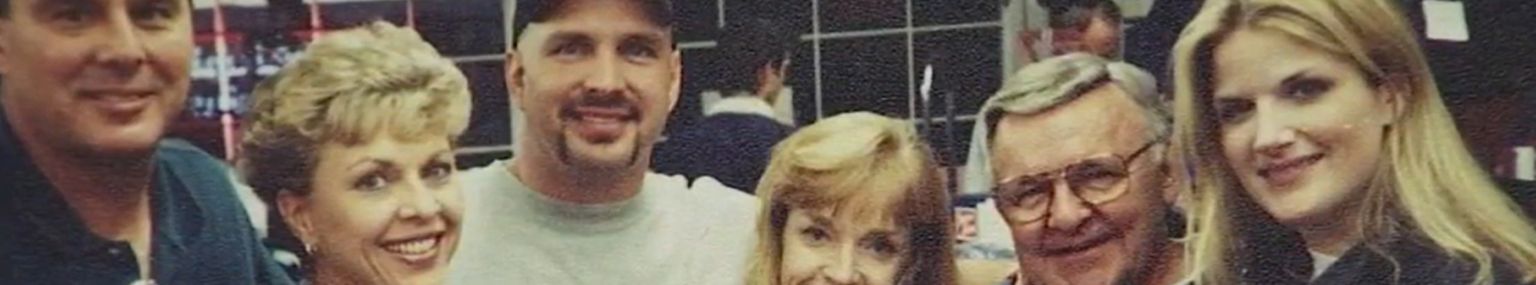 Six people stand closely together indoors, smiling at the camera. They appear to be posing for a group photo in a casual setting.