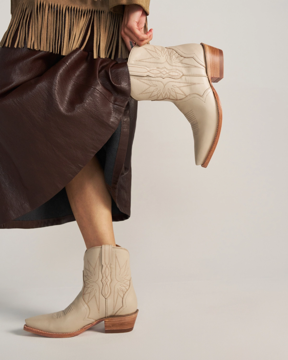 A person wearing a dark brown faux leather skirt, a tan fringe jacket, and light tan embroidered cowboy boots with wooden heels inspired by The Lynn Cowhide in a soft cream shade.