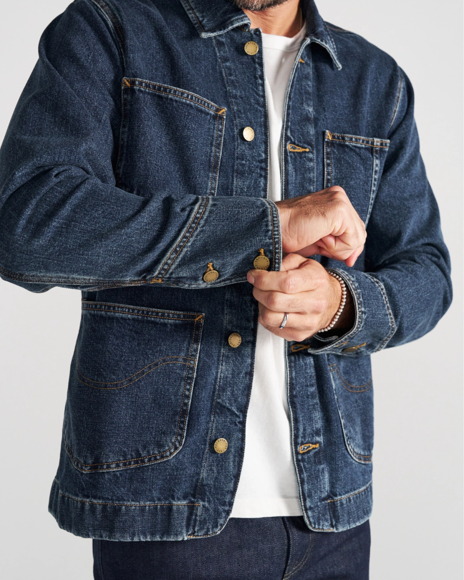 A person wearing a dark blue denim jacket over a white shirt fastens a button on the jacket sleeve.