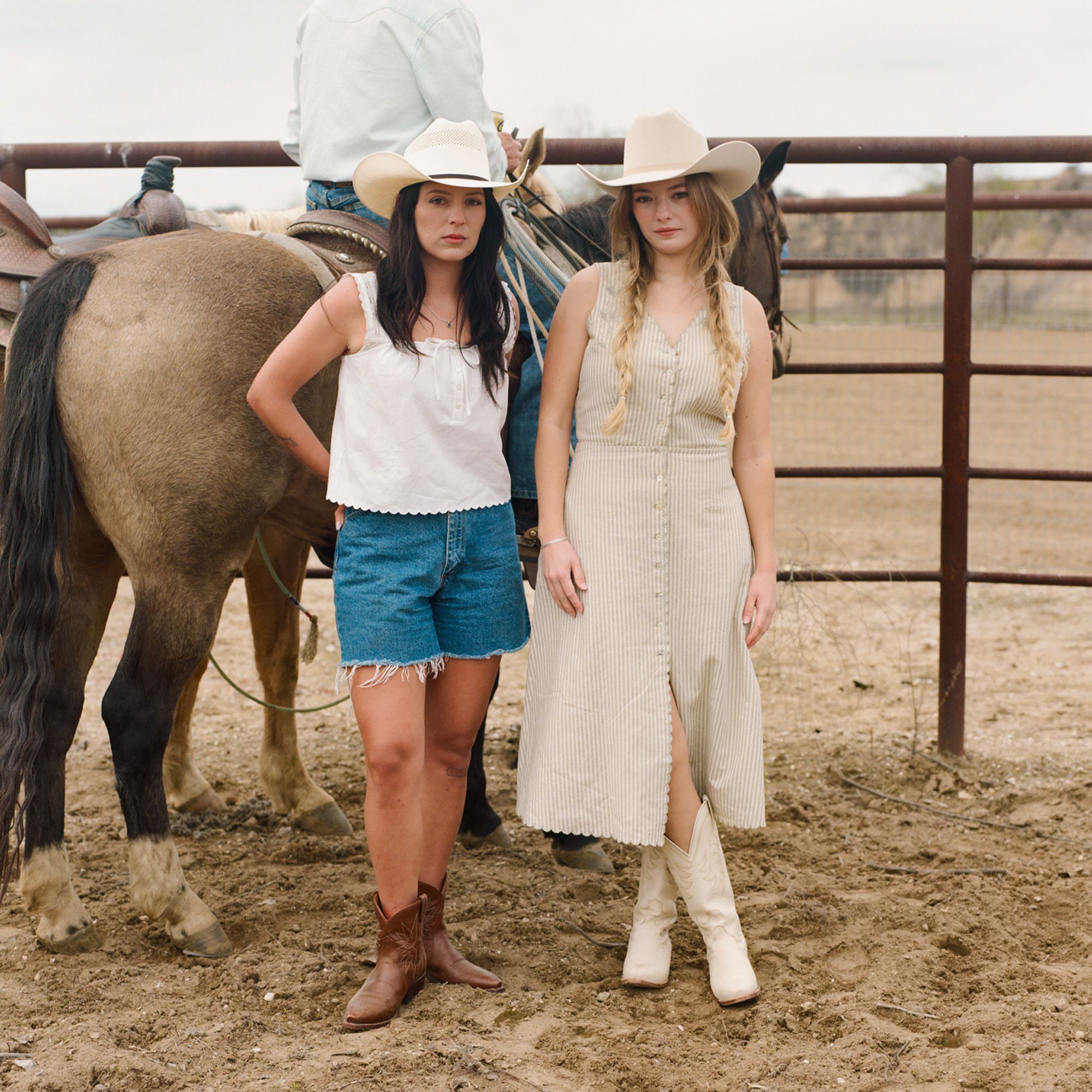 Two women in cowboy hats and boots stand next to a horse and a person on horseback, near a metal fence in a dirt corral area.