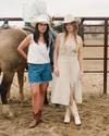 Two women in cowboy hats and boots stand next to a horse and a person on horseback, near a metal fence in a dirt corral area.