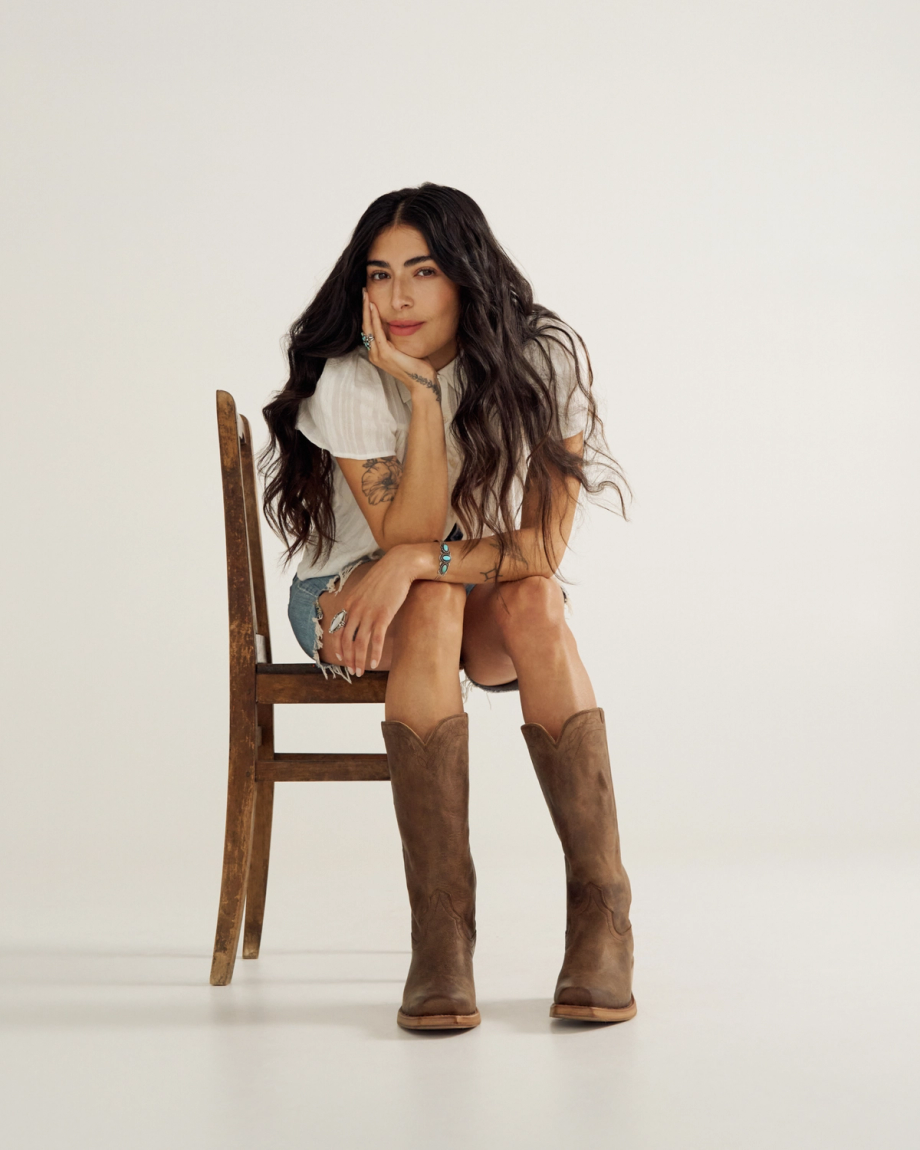 A woman with long dark hair sits on a wooden chair at The Charlie Goat cafe, resting her chin on her hand. She wears a white top, denim shorts, and brown cowboy boots against a plain background.