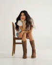 A woman with long dark hair sits on a wooden chair at The Charlie Goat cafe, resting her chin on her hand. She wears a white top, denim shorts, and brown cowboy boots against a plain background.