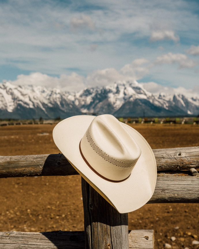 A cowboy hat rests on a wooden fence, with a snow-capped mountain range and a clear sky in the background.