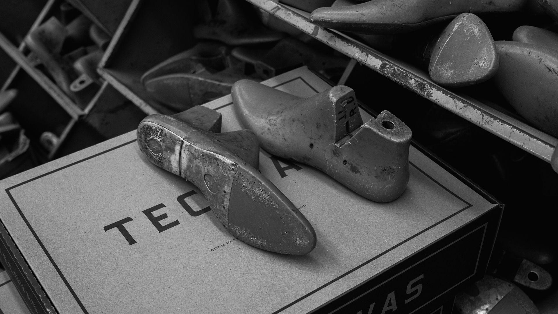 Two shoe lasts rest on a cardboard box labeled "TEC" among other shoe-making materials in a workshop setting.