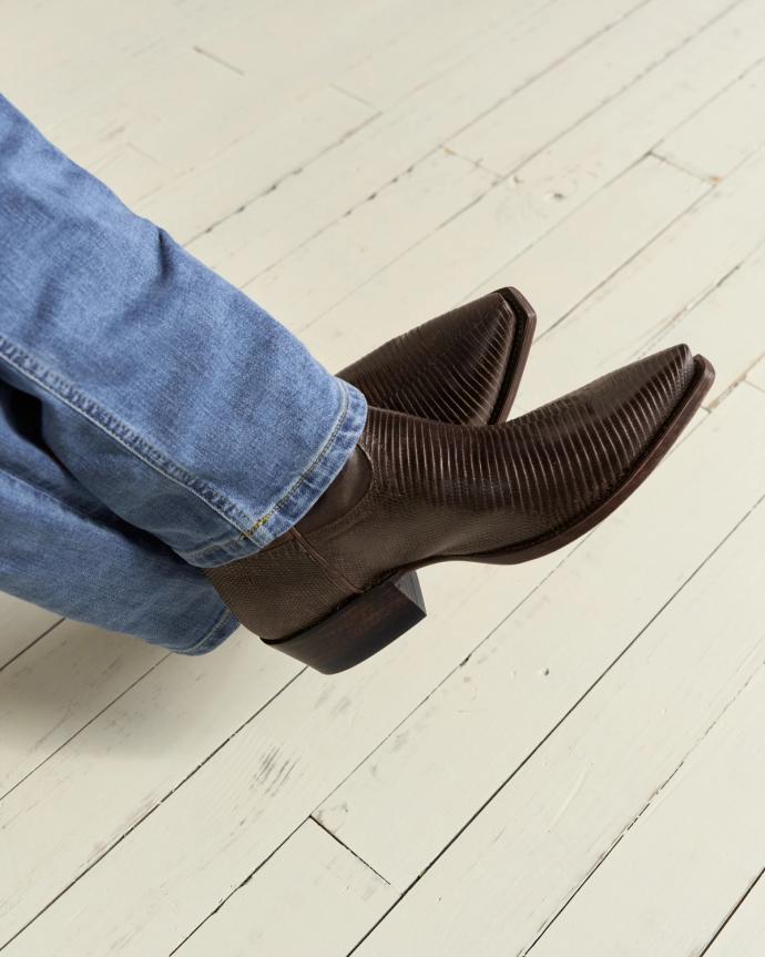 Person wearing brown cowboy boots and blue jeans, sitting on a light-colored wooden floor.