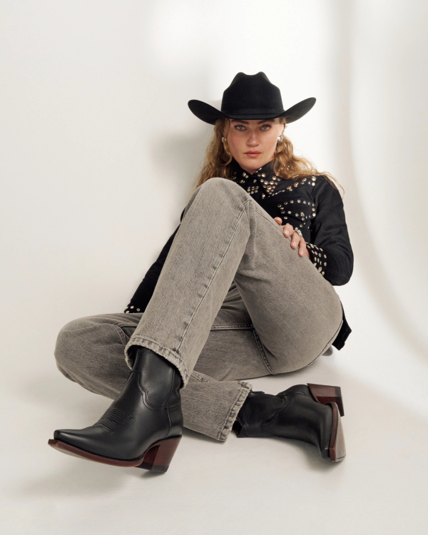 A person wearing a black cowboy hat, studded black jacket, grey jeans, and black boots sits on the floor against a plain white background.