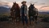 Cowgirl standing with two horses in front of a sunset