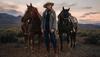 Cowgirl standing with two horses in front of a sunset