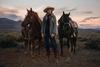 Cowgirl standing with two horses in front of a sunset