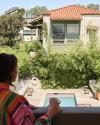 A person with a mug stands on a balcony overlooking a swimming pool and patio area surrounded by greenery and residential buildings.