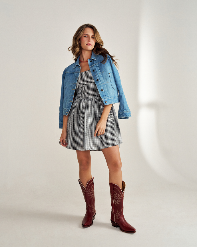 Person wearing a denim jacket, black and white checkered dress, and red cowboy boots stands against a plain background.