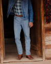 A person in a blue denim jacket and jeans, brown belt, plaid shirt, and brown cowboy boots stands in a wooden doorway. Red peppers hang on the right side of the doorway.