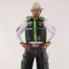 Man wearing a cowboy hat, protective vest, white shirt, and leather chaps stands with hands on hips against a plain background.