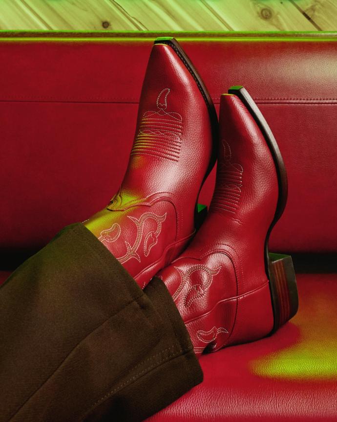Person wearing red leather cowboy boots with decorative stitching, resting feet on a red cushioned surface. The boots have pointed toes and wooden heels.