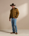 A man wearing a brown cowboy hat, brown jacket, blue jeans, and cowboy boots stands against a plain beige background.