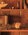 A lit candle with a cowboy boot design sits on a shelf with gold tinsel, surrounded by vintage and western-themed decor items.