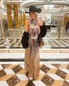 A woman in a stylish fringe dress and black hat stands in a luxurious lobby with marble floors and golden columns.