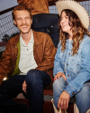 A man and woman, both smiling, are dressed in casual clothes. The man wears a brown jacket while the woman wears a denim jacket and a cowboy hat. They are seated outdoors, leaning against a fence.