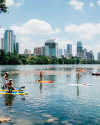 People are paddleboarding and kayaking on a calm river with a city skyline and high-rise buildings in the background.