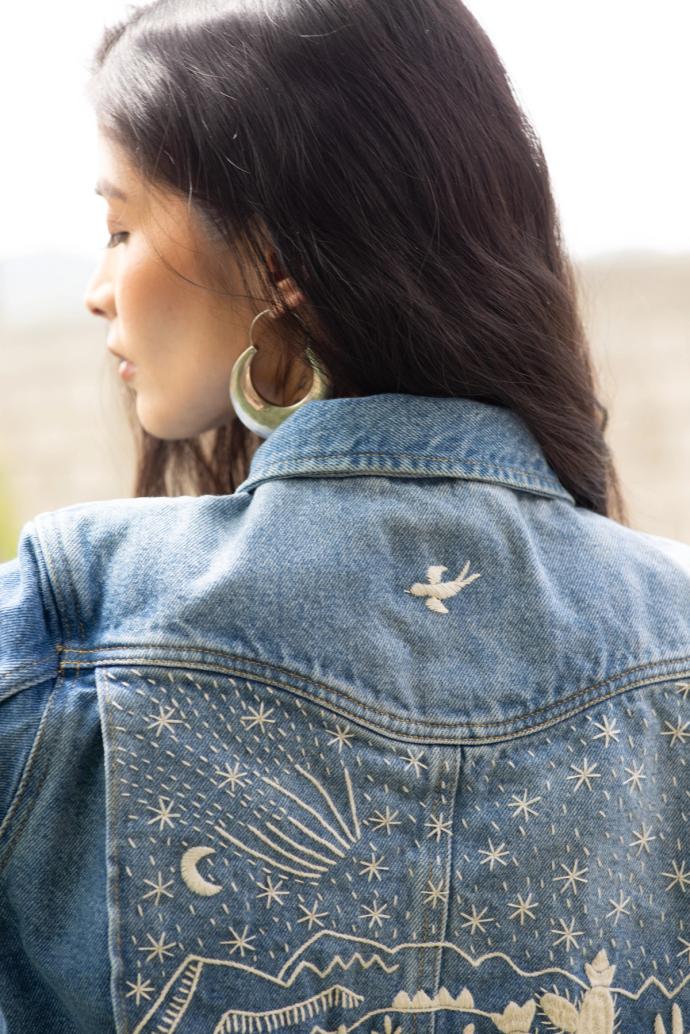 A woman with long dark hair faces away, showcasing her denim jacket adorned with embroidered designs of stars, a moon, a shooting star, and a bird. She is wearing large hoop earrings.