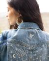 A woman with long dark hair faces away, showcasing her denim jacket adorned with embroidered designs of stars, a moon, a shooting star, and a bird. She is wearing large hoop earrings.