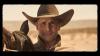 A man in a cowboy hat stands next to a horse in a desert landscape, looking directly at the camera.