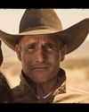 A man in a cowboy hat stands next to a horse in a desert landscape, looking directly at the camera.